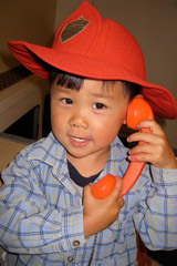 A boy wearing a firemen's hat holding a toy phone up to his ear.