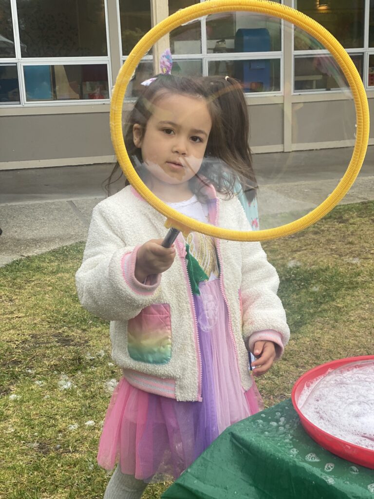 A young girl holding up a giant bubble blowing ring, looking through the center of the bubble